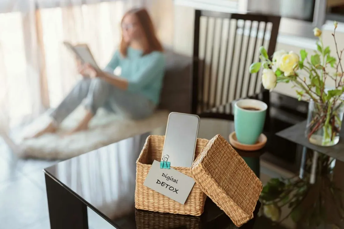 Smartphone in einem Korb mit „Digital Detox“-Schild, im Hintergrund liest eine Frau entspannt ein Buch